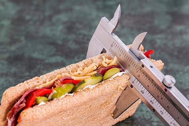 Een metalen schuifschaal meet de dikte van de sandwich. De sandwich wordt in plakjes gesneden en belegd met komkommers, rode paprika, salami en geraspte kaas.